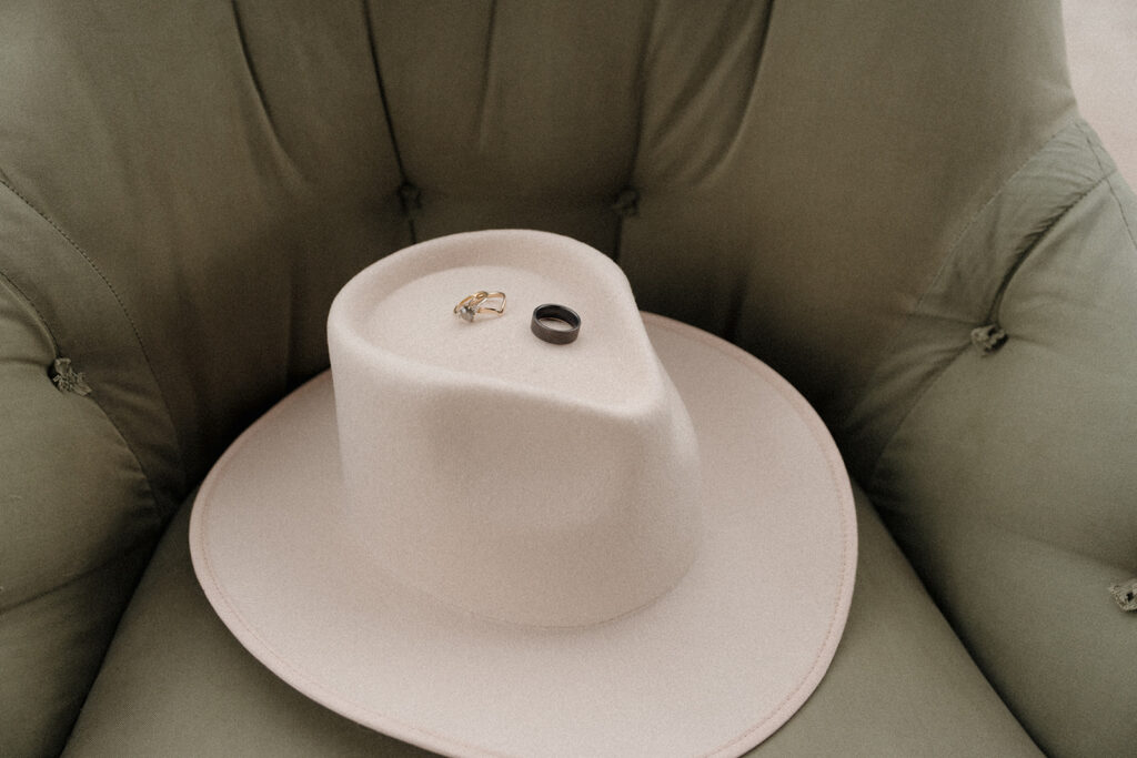cream color cowboy hat sitting on a green chair with bride and grooms rings